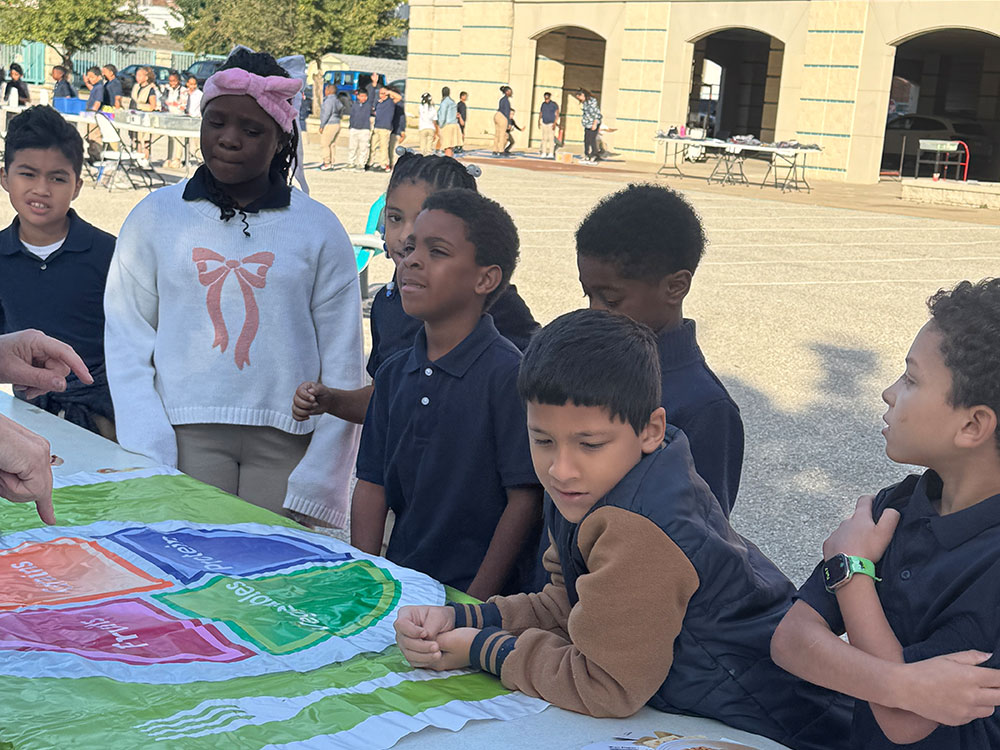 A group of children in a schoolyard gather around a colorful educational board, focused and engaged. A teacher's hand points to a section.