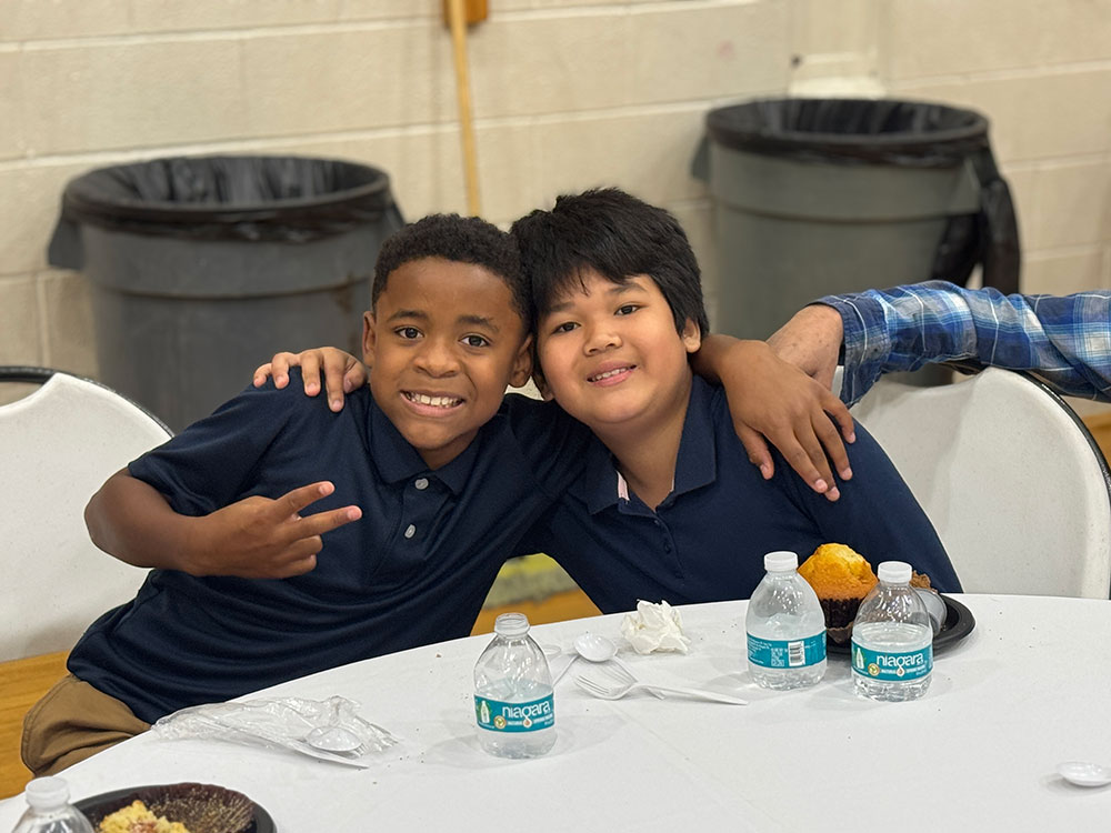 Two boys smiling and sitting at a table, embracing each other. One is making a peace sign. Water bottles and plates with food are on the table.