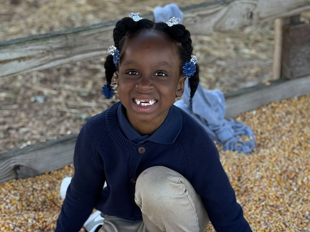 A smiling child with neatly braided hair sits on a pile of corn kernels. They wear a blue sweater and beige pants, exuding joy and playfulness.