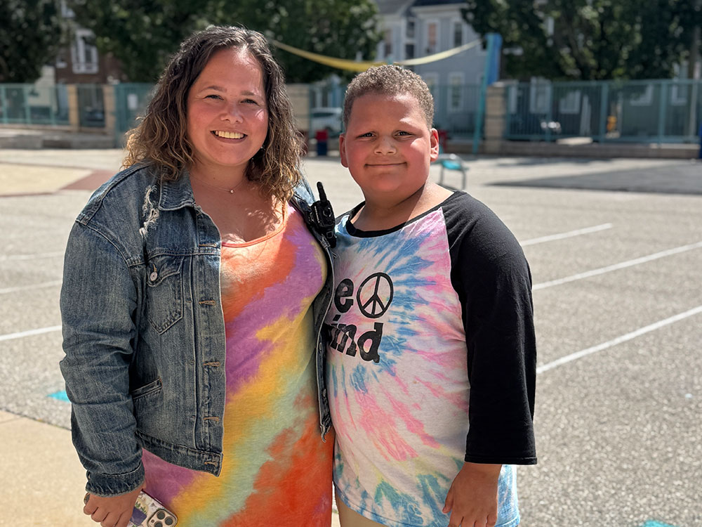 A smiling woman and boy stand in a sunny playground. She wears a denim jacket and colorful dress; he wears a tie-dye shirt with "Be Kind."